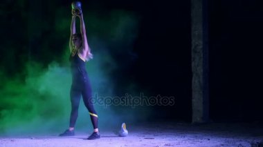 Ağırlıkları ile çeşitli güç egzersizleri yapıyor atletik, güzel genç bir kadın. Gece, terk edilmiş eski bir hangarda hafif duman, siste çok renkli projektörler ışığında,