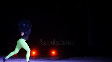 Atletik, genç kadının bir halter, lunges, ile çeşitli güç egzersizleri yapması ağız kavgası. Gece, terk edilmiş eski bir hangarda hafif duman, siste çok renkli projektörler ışığında,