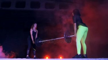 İki atletik kızlar bir halter ile çeşitli güç egzersizleri yapıyor, ağız kavgası, bir eski terk edilmiş hangarda hafif duman, siste çok renkli projektörler ışığında geceleri bina