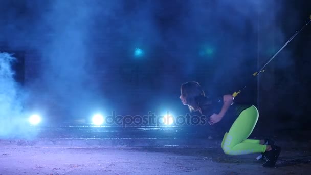 Athletic, sexy woman performs exercises with fitness trx system, TRX suspension straps. At night, in light smoke, fog, in light of multicolored searchlights, in abandoned building — Stock Video