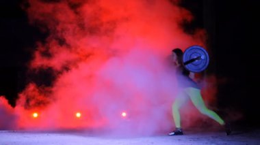 Atletik, genç kadının bir halter, lunges, ile çeşitli güç egzersizleri yapması ağız kavgası. Gece, terk edilmiş eski bir hangarda hafif duman, siste çok renkli projektörler ışığında,