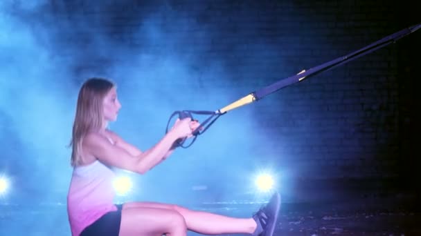 Athletic, sexy woman performs exercises with fitness trx system, TRX suspension straps. At night, in light smoke, fog, in light of multicolored searchlights, in abandoned building — Stock Video