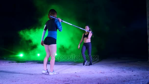 Twee sportieve meisjes in paar doen verschillende sterkte oefeningen met een barbell, squats, 's nachts, in lichte rook, mist, in het licht van veelkleurige zoeklichten, in een oude verlaten hangar, — Stockvideo