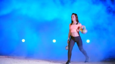 Ağırlıkları ile çeşitli güç egzersizleri yapıyor atletik, güzel genç bir kadın. Geceleri hafif duman, sis, çok renkli projektörler ışığında içinde binayı terk edilmiş