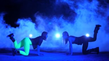 İki atletik kadın, kendi ayakları üzerinde gece, hafif duman, sis, bir eski terk edilmiş hangarda çok renkli projektörler ışığında ağırlık ölçme ile fitness egzersizleri yapıyor,