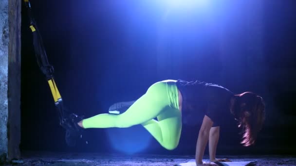 Athlétique, femme sexy effectue des exercices avec le système de fitness trx, sangles de suspension TRX. La nuit, dans la fumée légère, le brouillard, à la lumière de projecteurs multicolores, dans un bâtiment abandonné — Video
