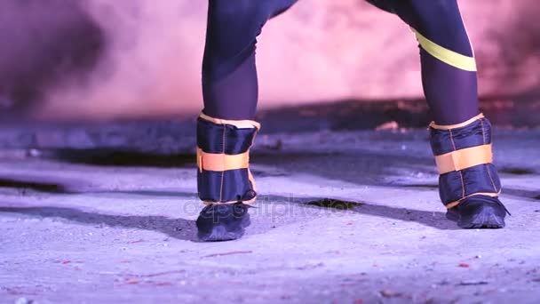 Close-up van de benen van de twee vrouwen met wegingen, doen van fitness oefeningen, 's nachts, in lichte rook, mist, in het licht van veelkleurige zoeklichten, in een oude verlaten hangar, — Stockvideo