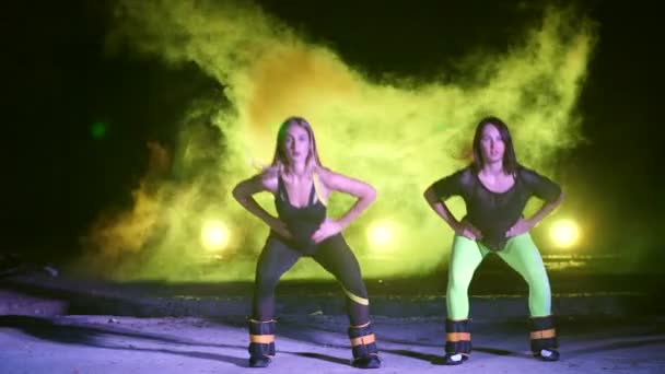 Twee sportieve vrouwen, doen van verschillende fitness oefeningen met weging op hun benen, 's nachts, in lichte rook, mist, in het licht van veelkleurige zoeklichten, in een oude verlaten hangar, — Stockvideo