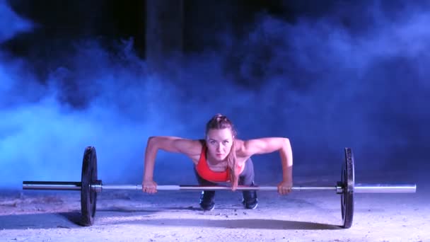 Atletisk flicka gör push ups med en skivstång, på natten, i ljus rök, dimma, mot bakgrund av mångfärgade strålkastare, i en gammal övergiven hangar, bygga — Stockvideo