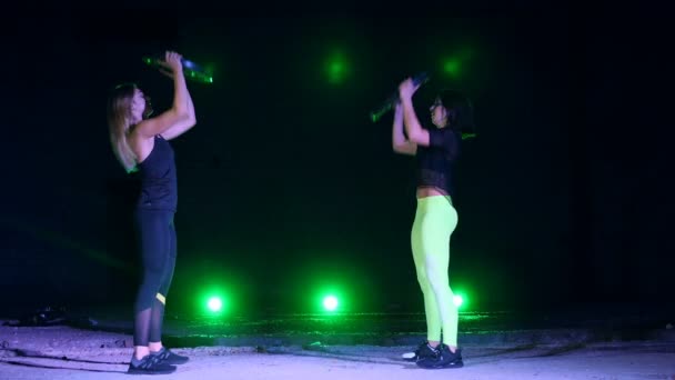 Duas atléticas, bonitas, mulheres fazendo exercícios de força com placas pesadas, à noite, em fumaça leve, neblina, à luz de holofotes multicoloridos, em um antigo hangar abandonado , — Vídeo de Stock