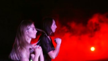 İki atletik kadın fitness egzersizleri yapıyor. Gece, terk edilmiş eski bir hangarda çok renkli projektörler ışığında hafif duman, sis,