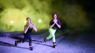 İki atletik kadın, kendi ayakları üzerinde gece, hafif duman, sis, bir eski terk edilmiş hangarda çok renkli projektörler ışığında ağırlık ölçme ile çeşitli fitness egzersizleri yapıyor,