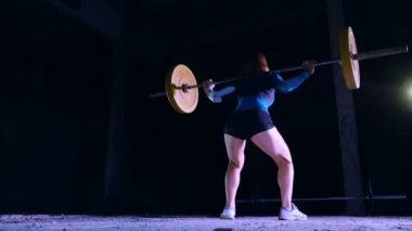 atletik genç kadın, halter yükseltir, halter ile mekik yok. Arkadan Görünüm. Geceleri projektörler, hafif duman, sis, bir eski terk edilmiş hangarda ışığında bina.