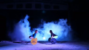 İki atletik kızlar, sporcular, halter yükseltmek, mekik halter ile yap. Geceleri, projektörler, hangarda bir eski terk edilmiş, Bina hafif duman, sis ışığında.