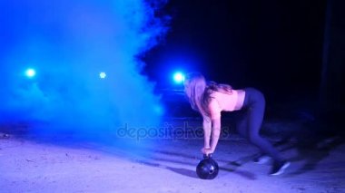 Ağırlıkları ile çeşitli güç egzersizleri yapıyor atletik, güzel genç bir kadın. Geceleri, projektörler, hangarda bir eski terk edilmiş, Bina hafif duman, sis ışığında