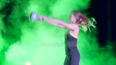 Ağırlıkları ile çeşitli güç egzersizleri yapıyor atletik, güzel genç bir kadın. Geceleri hafif duman, sis, çok renkli projektörler ışığında içinde binayı terk edilmiş
