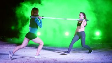Çift bir halter, lunges, ağız kavgası, gece, terk edilmiş eski bir hangarda çok renkli projektörler ışığında hafif duman, sis ile çeşitli güç egzersizleri yapıyor iki atletik kızlar,