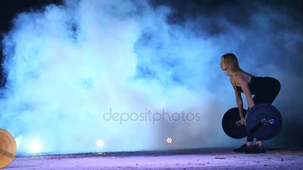 Спортивна молода жінка, піднімає штангу, сідає з штангою. Вночі, у світлі прожекторів, у світлому димі, тумані, у старому покинутому ангарі, будівлі — стокове відео