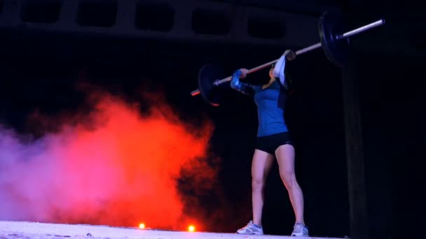Atletisk, ung kvinna väcker barbell ovanför huvudet. På natten, mot bakgrund av mångfärgade strålkastare, i ljus rök, dimma, i en gammal övergiven hangar, — Stockvideo