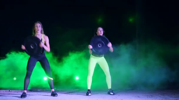 Dos mujeres atléticas, hermosas, haciendo ejercicios de fuerza con placas pesadas, por la noche, en humo ligero, niebla, a la luz de reflectores multicolores, en un viejo hangar abandonado , — Vídeos de Stock