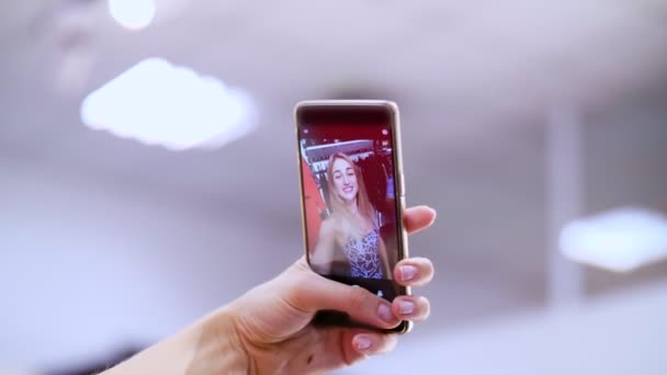 Chica sexy, una hermosa mujer rubia hace selfie en un traje nuevo, sonriendo, en una tienda, una boutique de ropa. En el marco hay una mano femenina con un teléfono inteligente. cámara lenta — Vídeos de Stock