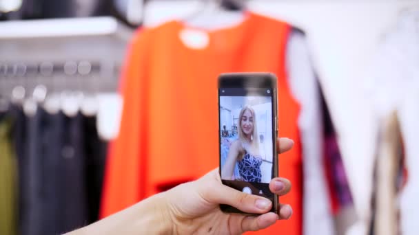 Chica sexy, una hermosa mujer rubia hace selfie en un traje nuevo, sonriendo, en una tienda, una boutique de ropa. En el marco hay una mano femenina con un teléfono inteligente. cámara lenta — Vídeos de Stock