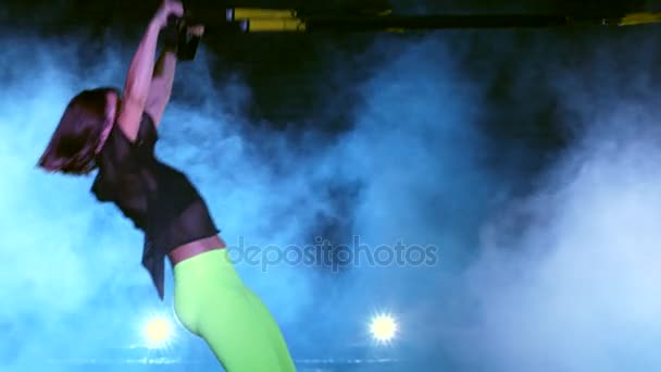 Атлетичная, сексуальная женщина выполняет упражнения с системой фитнес-трекс, ремнями подвески TRX. Ночью, в легком дыму, тумане, в свете разноцветных прожекторов, в заброшенном здании — стоковое видео