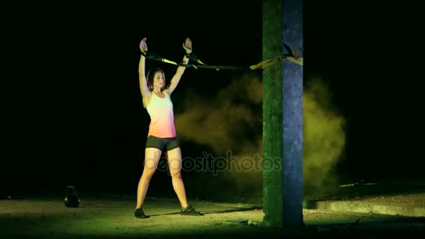 Atlético, mulher sexy executa exercícios com sistema de fitness trx, cintas de suspensão TRX. À noite, em fumaça leve, neblina, à luz de holofotes multicoloridos, em prédio abandonado — Vídeo de Stock