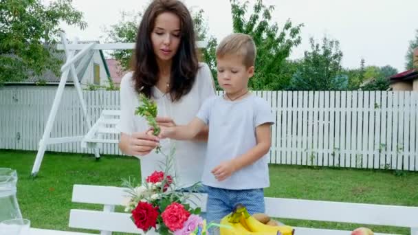 Летом, в саду. Мама с четырехлетним сыном делает букет цветов. Мальчику это очень нравится, он счастлив, веселится, семья проводит досуг вместе . — стоковое видео