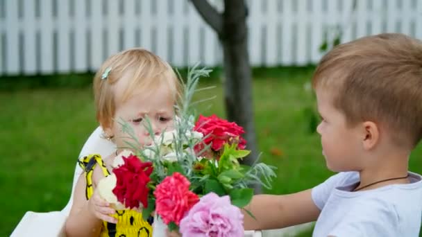 Zomer in de tuin. de vier-jaar-oude jongen geeft een boeket bloemen aan zijn jongere zus van de een-jaar-oude het meisje houdt van bloemen, ze is ze, het meisje eet een appel. — Stockvideo