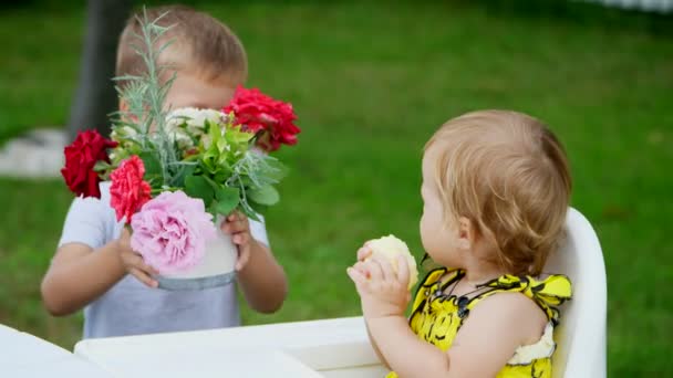 Verano, en el jardín. el niño de cuatro años le da un ramo de flores a su hermana menor de un año, hermano besa a su hermana en la mejilla. La chica come una manzana — Vídeos de Stock