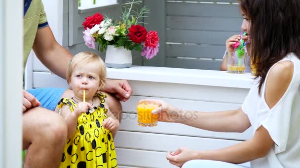 Sommaren i trädgården, föräldrar spela med små barn, en flicka och en pojke, på ett café, i barnens lekstuga, behandla barn med färskpressad juice, dricka juicer. — Stockvideo