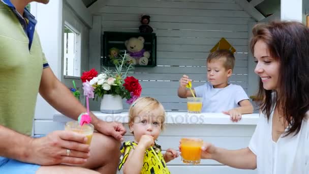 Летом в саду родители играют с маленькими детьми, девочкой и мальчиком, в кафе, в детском домике, угощают детей свежевыжатыми фруктовыми соками, пьют соки . — стоковое видео