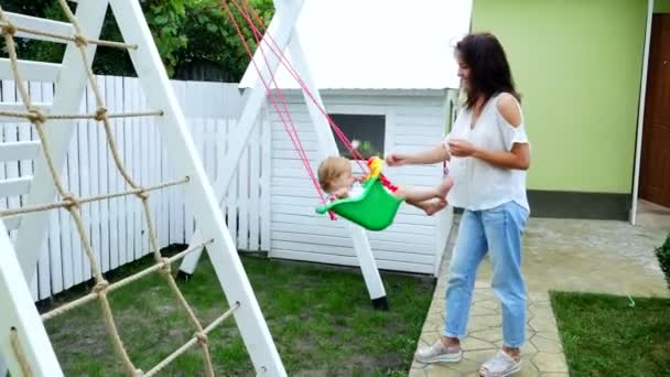Été, dans le jardin, mère, belle jeune femme, balançant sur une balançoire sa fille d'un an. La famille passe son temps libre ensemble . — Video