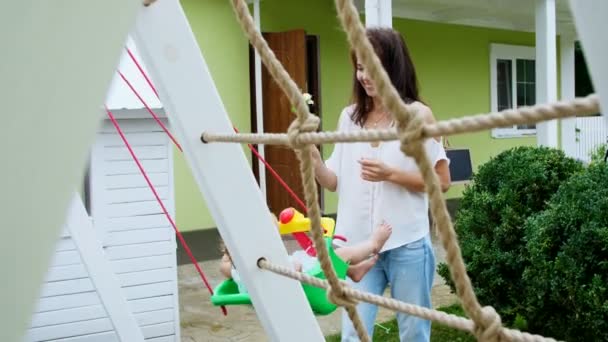 Verano, en el jardín, madre, hermosa joven, balanceándose en un columpio de su hija de un año de edad. La familia pasa su tiempo libre juntos. . — Vídeo de stock