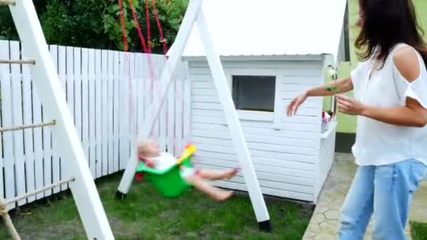 Sommer, im Garten, Mutter, schöne junge Frau, schwingt auf einer Schaukel ihre einjährige Tochter. die Familie verbringt ihre Freizeit zusammen. — Stockvideo