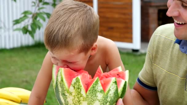 在夏天，在花园里，父亲带着四岁的儿子伤一个西瓜，吃它，玩得开心，一个小男孩非常喜欢西瓜。甜甜的西瓜，和家人一起吃午饭. — 图库视频影像