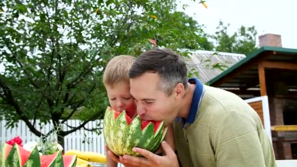 En verano, en el jardín, padre con un hijo de cuatro años de edad, cortar una sandía y comer, divertirse, un niño le gusta mucho la sandía. sandía dulce para el almuerzo con la familia . — Vídeos de Stock