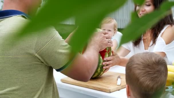 Mladá rodina se dvěma malými dětmi legrační sedí u jídelního stolu v zahradě, v létě. Táta škrty velký meloun, se bude považovat jeho rodinu. oběd s rodinou.