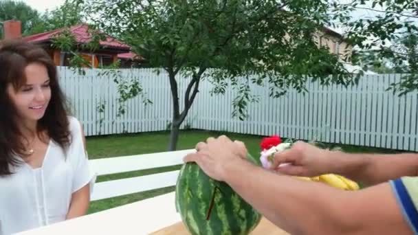 Een jong gezin met twee kleine grappige kinderen zitten op een tafel in de tuin, in de zomer. Papa snijdt een grote watermeloen, gaat voor de behandeling van zijn familie. lunch met de familie. — Stockvideo