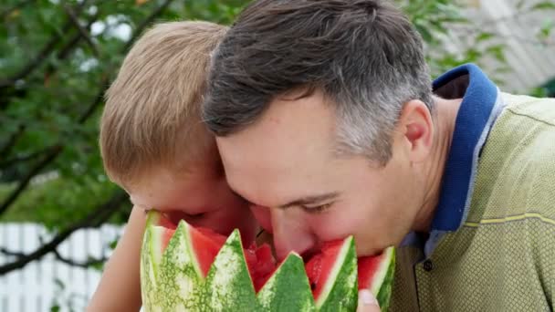 En verano, en el jardín, padre con un hijo de cuatro años de edad, cortar una sandía y comer, divertirse, un niño le gusta mucho la sandía. sandía dulce para el almuerzo con la familia . — Vídeos de Stock