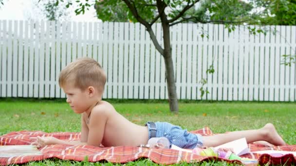 Чотирирічний хлопчик читає книгу, в саду, лежачи на ковдрі, ковдрі, на траві, газоні, влітку — стокове відео