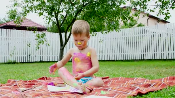 Uma criança pequena, um menino de quatro anos brincando, pintando com pinturas de dedo, decorando-se, no jardim, sentado em um cobertor, na grama, gramado, no verão. ele se divertindo — Vídeo de Stock