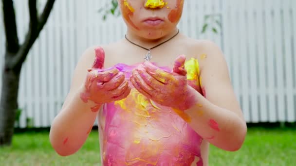 Un niño pequeño, un niño de cuatro años jugando, pintando con pinturas de dedos, decorándose a sí mismo, en el jardín, sentado en una manta, sobre hierba, césped, en el verano. Él se divierte — Vídeo de stock