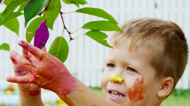 Uma criança, um menino de quatro anos brincando, pintando com pinturas de dedo, decorando folhas nas árvores do jardim, no verão . — Vídeo de Stock