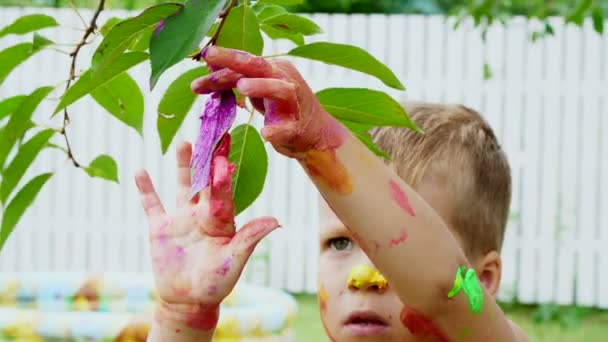 Ein kleines Kind, ein vierjähriger Junge beim Spielen, Malen mit Fingerfarben, Dekorieren der Blätter an den Bäumen im Garten, im Sommer. — Stockvideo