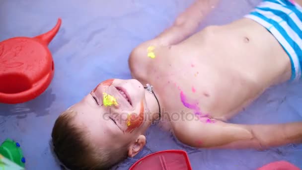 Ein vierjähriger Junge liegt lächelnd in einem aufblasbaren Kinderbecken, planscht Wasser. in der Nähe schwimmt Spielzeug. Blick von oben. — Stockvideo