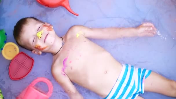 En fyra-årig pojke ligger i en uppblåsbar barnpool, stänkande vatten, leende. I närheten float leksaker. Visa från ovan. — Stockvideo