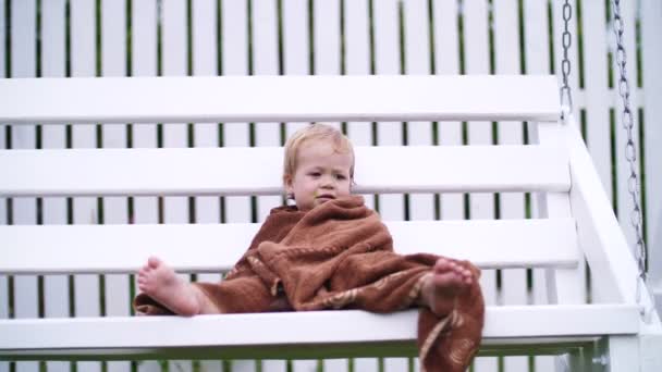 Una niña de un año, envuelta en una toalla, con una cara sucia, sola sentada en un columpio en el jardín, en el verano. Tiene una mirada triste. Ella quiere dormir. — Vídeos de Stock