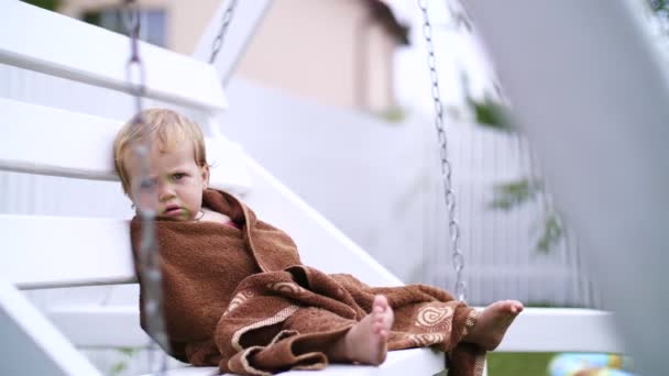 Una niña de un año, envuelta en una toalla, con una cara sucia, sola sentada en un columpio en el jardín, en el verano. Tiene una mirada triste. Ella quiere dormir. — Vídeos de Stock